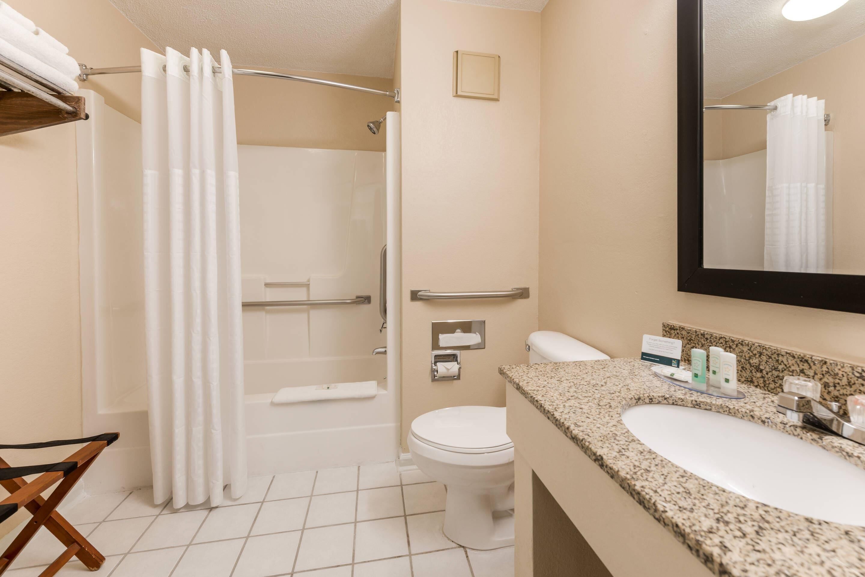 Un baño con ducha, inodoro y lavamanos, equipado con barras de apoyo y un espejo.