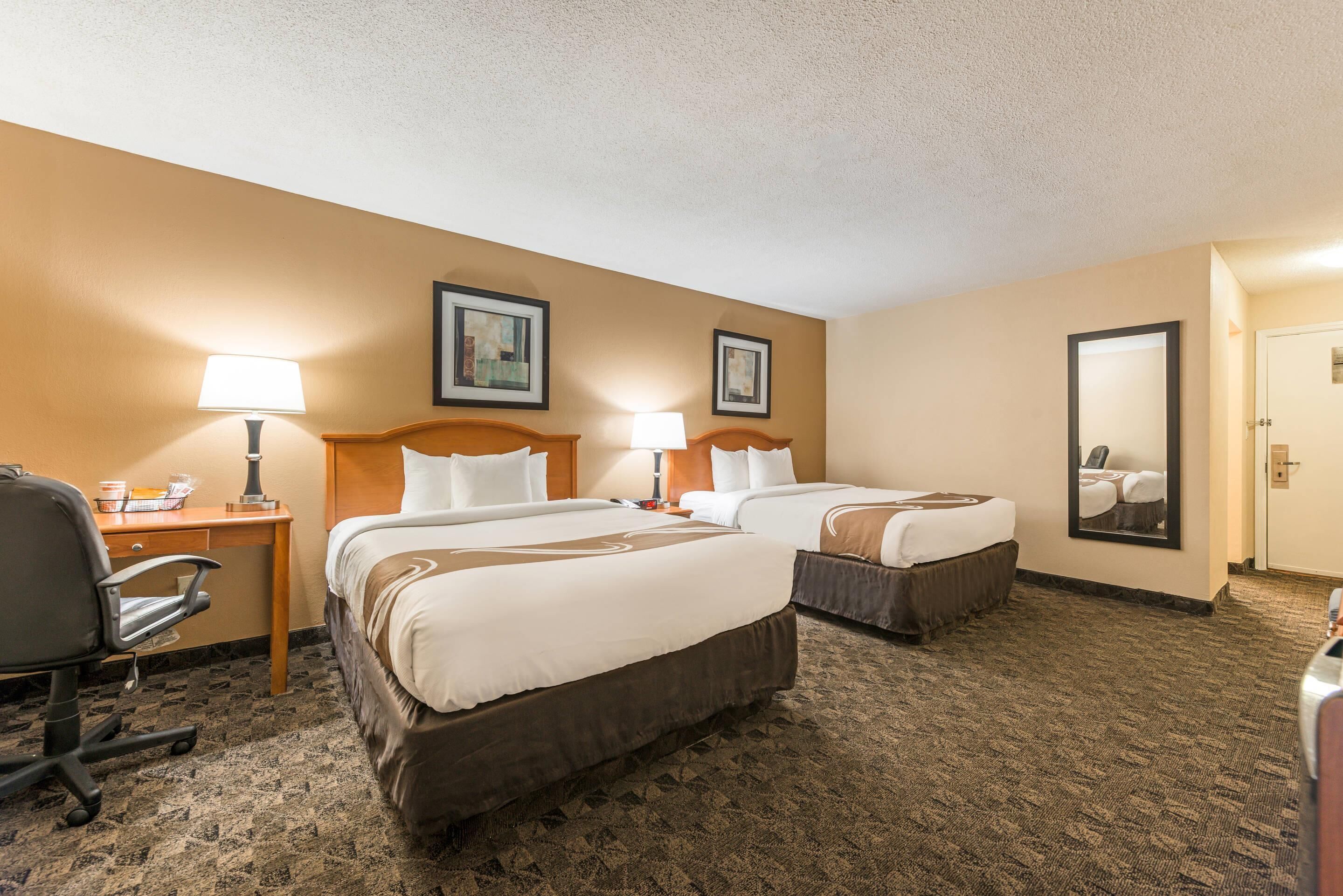 A hotel room with two beds, a desk with a lamp, a chair, and a carpeted floor.