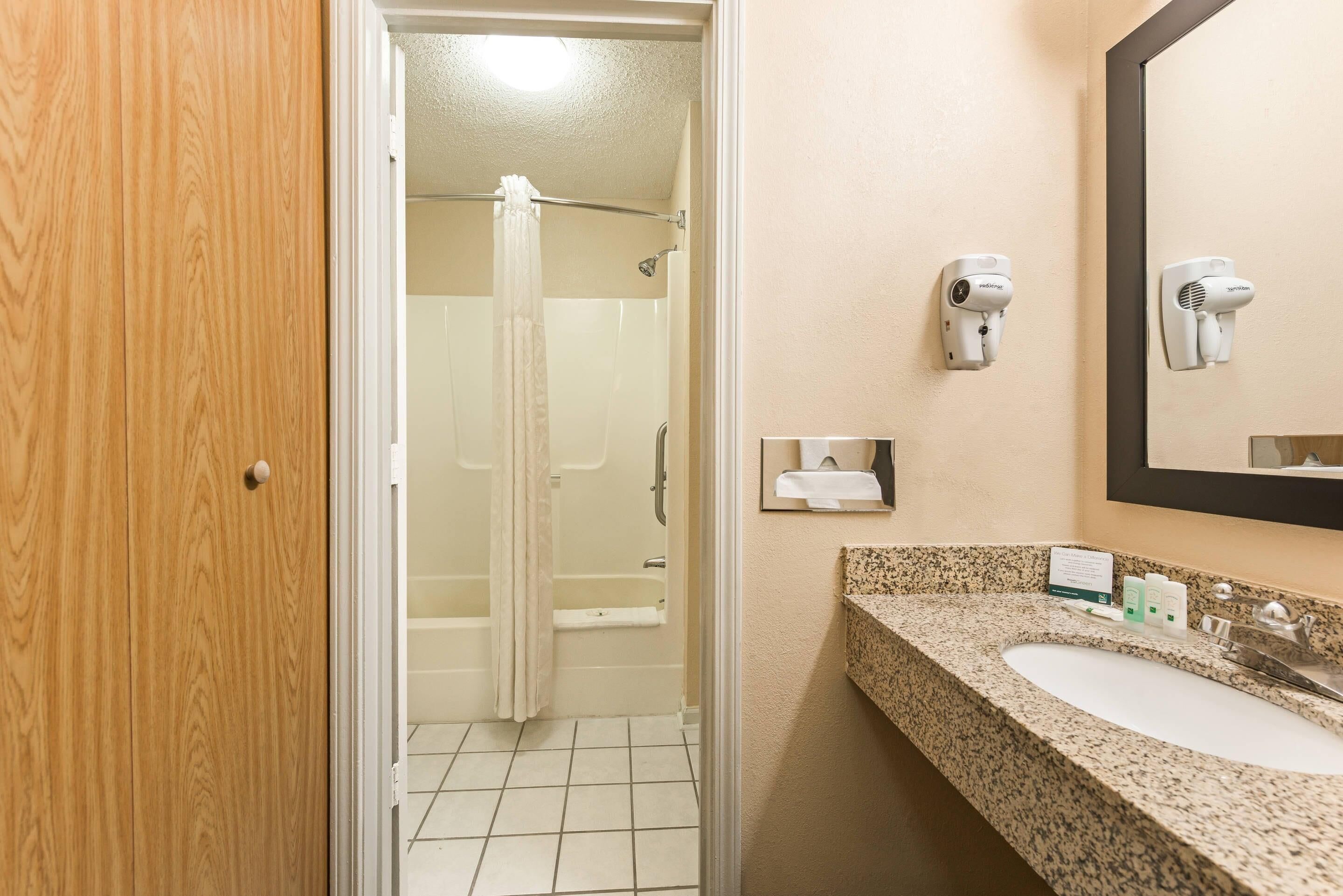 A bathroom with a wooden door, a shower, a sink with a granite countertop, and two hair dryers mounted on the wall.