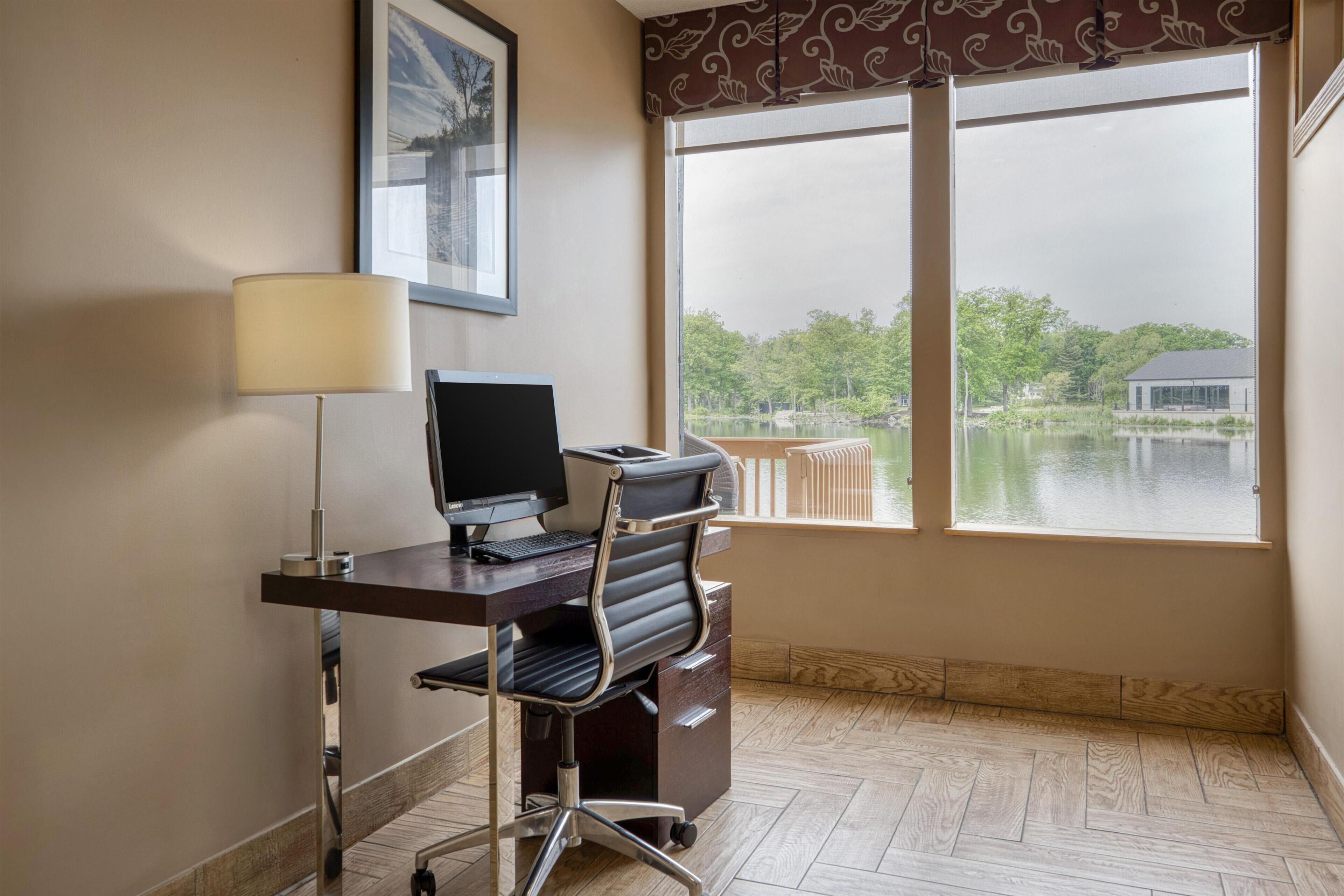 A room with a desk, computer, chair, lamp, and a large window with a view of a lake and buildings.