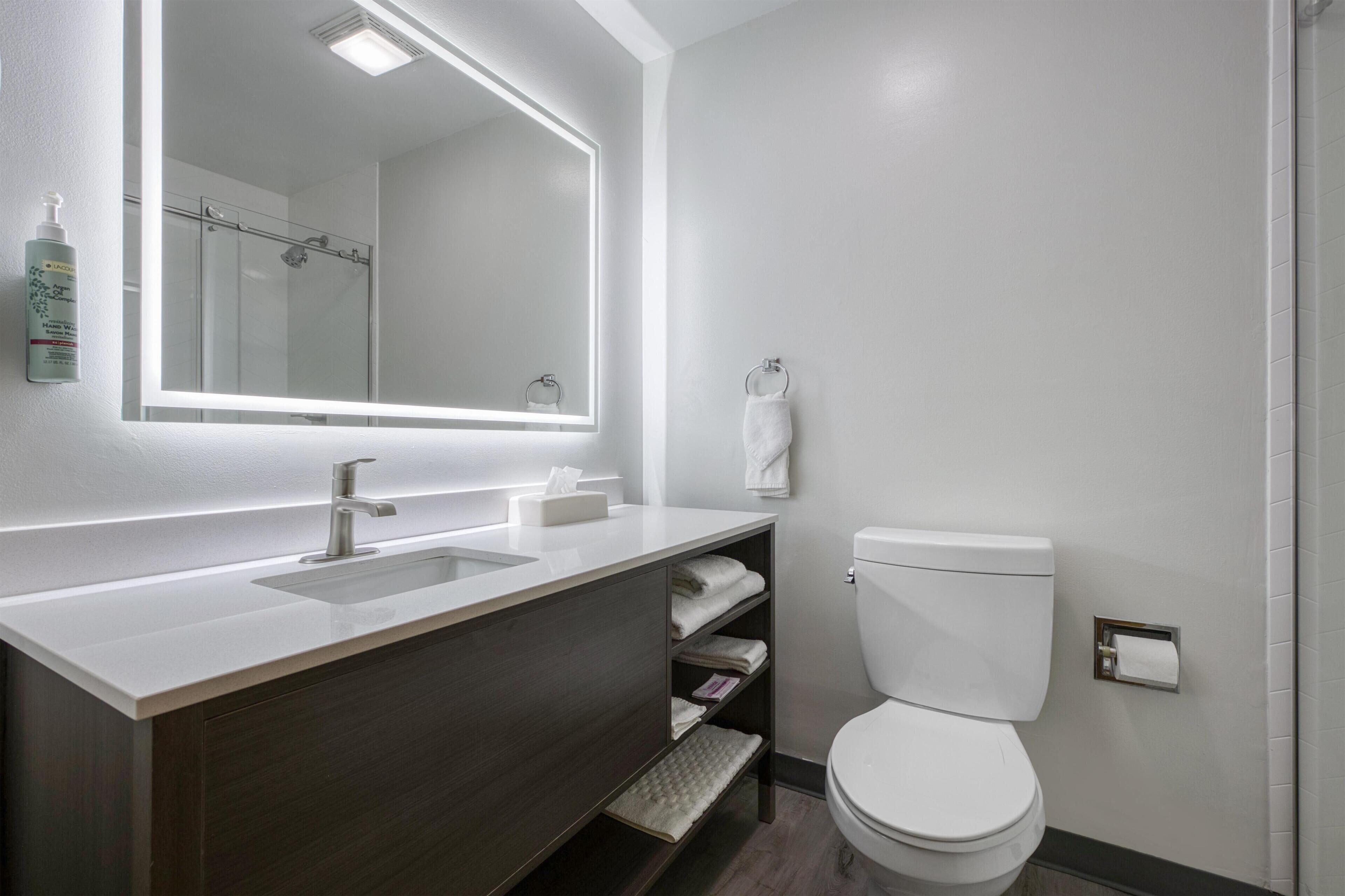 Un baño moderno con un lavamanos blanco, tocador de madera oscura, un inodoro y un espejo grande con iluminación integrada.