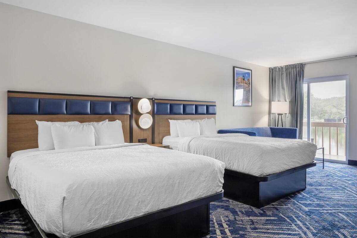 A hotel room with two beds, a large window with a view, a blue carpet, and a headboard with a clock.