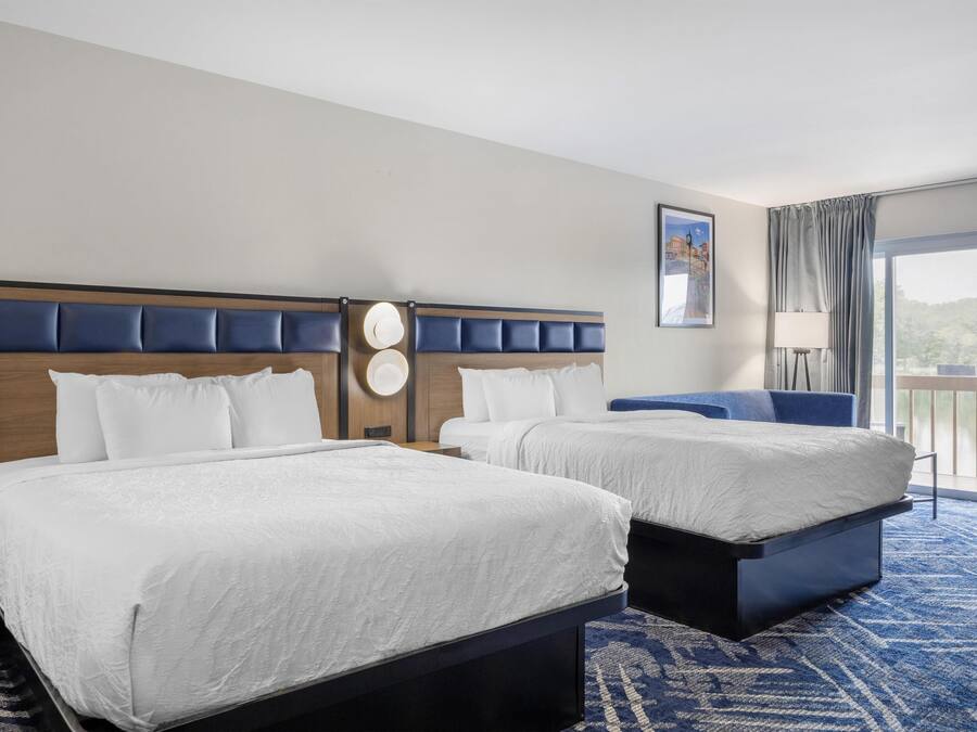 Une chambre d’hôtel avec deux lits, une grande fenêtre donnant sur un beau paysage, une moquette bleue et une tête de lit ornée d’une horloge.