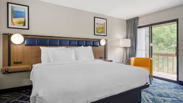 Habitación de hotel con una cama grande, una mesita de noche, una silla, un ventanal con vistas y dos cuadros en la pared.