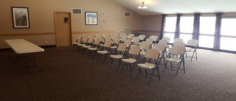 A conference room with a long table and rows of chairs, large windows with curtains, and a door.