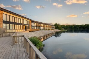 Un bâtiment moderne doté de grandes fenêtres et de balcons, d’une terrasse en bois et donnant sur un lac paisible.