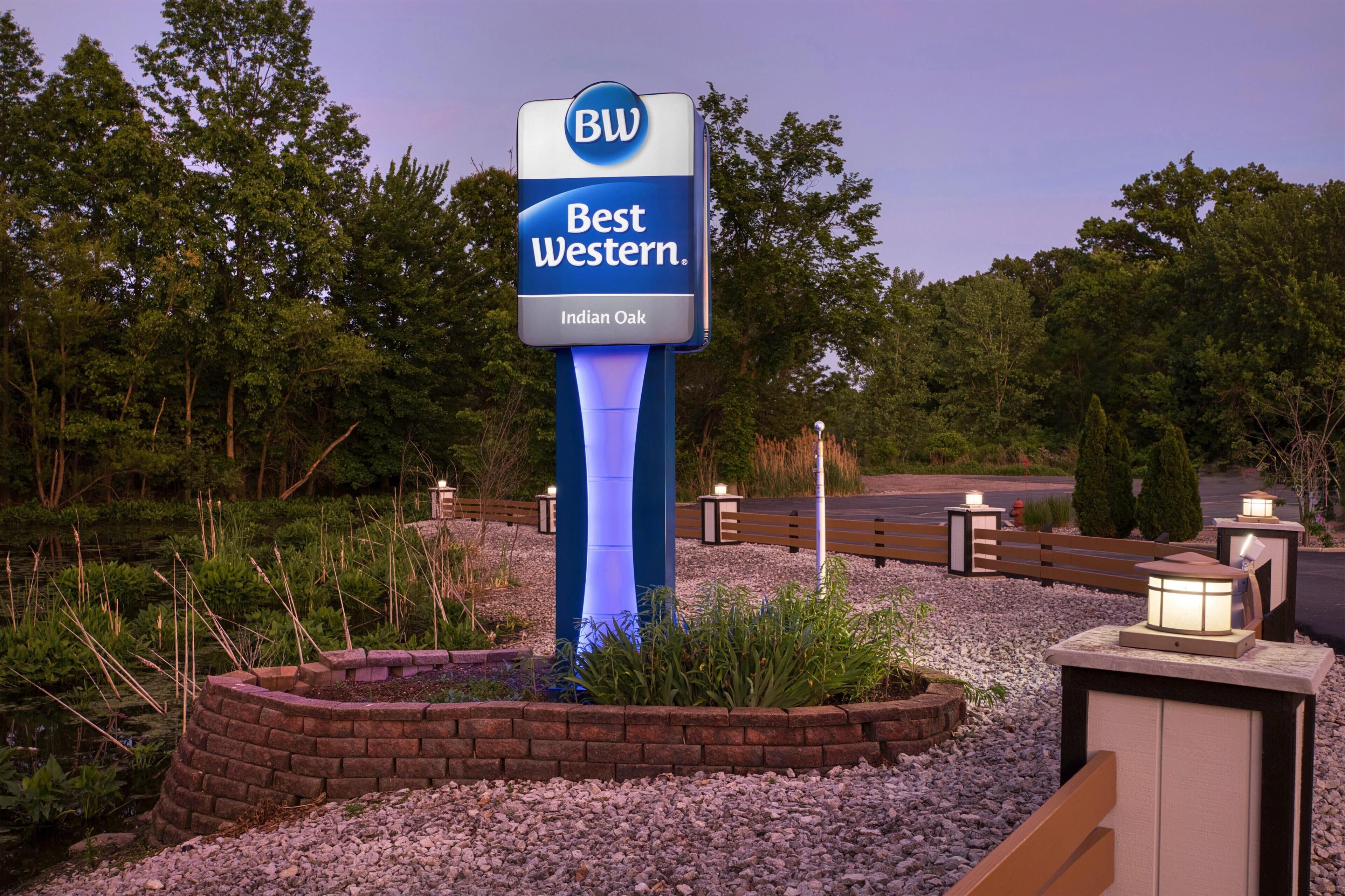 A Best Western hotel sign with a blue and white color scheme, surrounded by a landscaped area with a pond, benches, and decorative lighting.