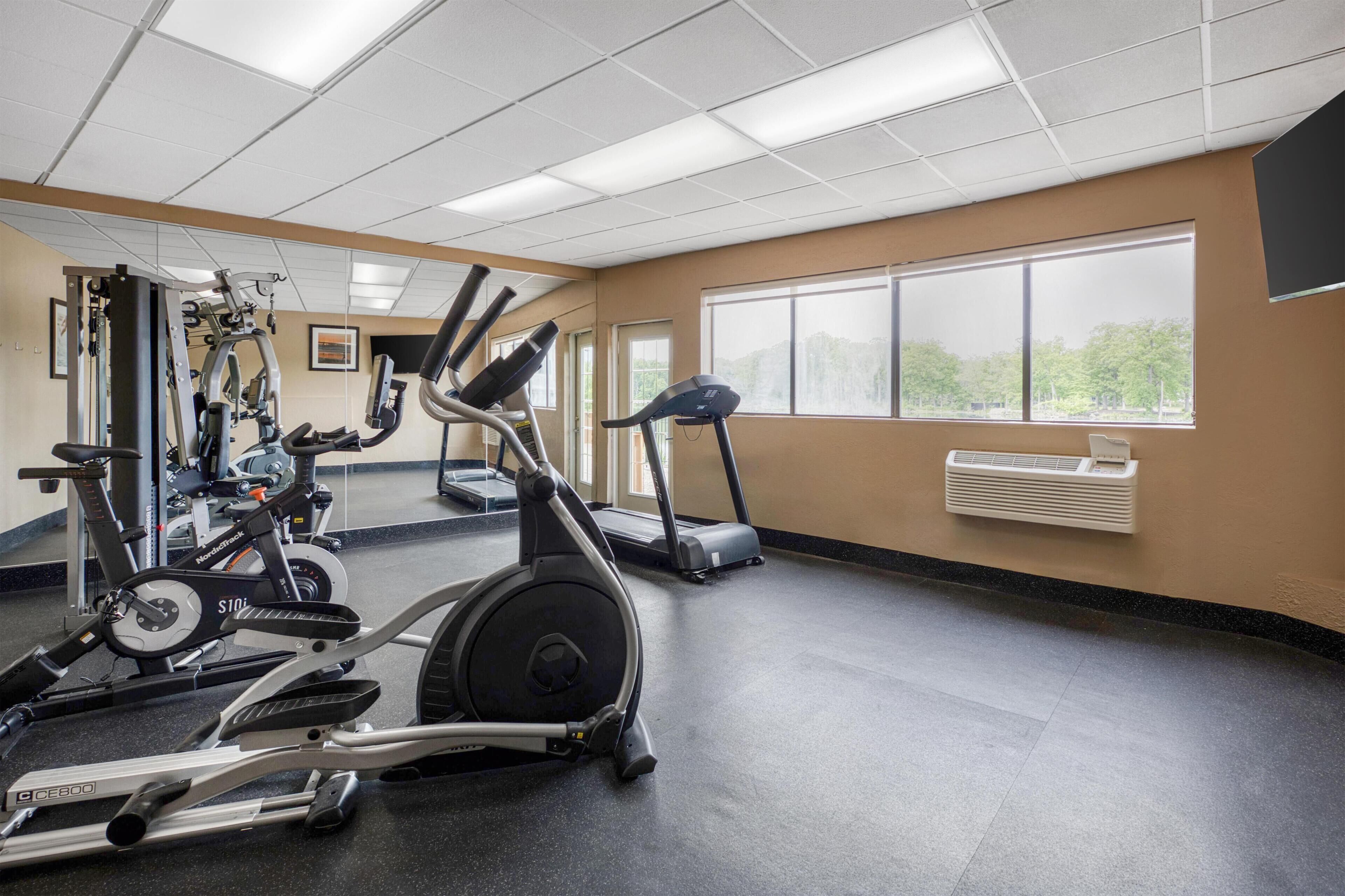 A gym with cardio machines, a mirrored wall, and large windows.