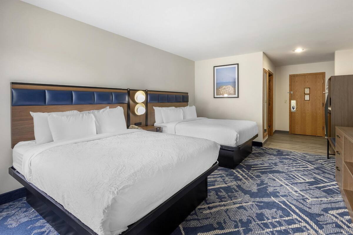 A hotel room with two beds, a wooden headboard, a patterned rug, and a framed picture on the wall.