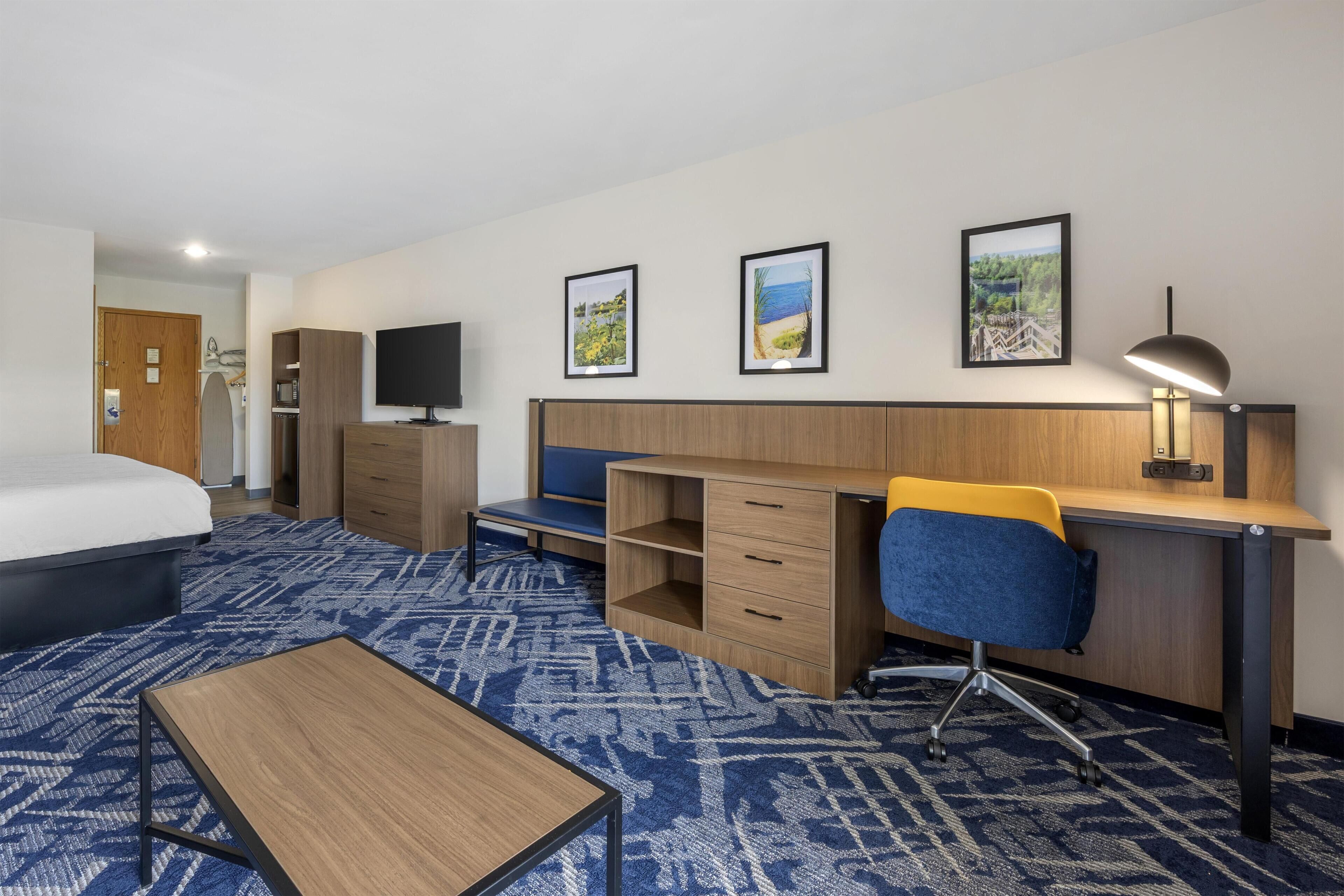 A hotel room with a bed, desk, chair, TV, and artwork on the wall.
