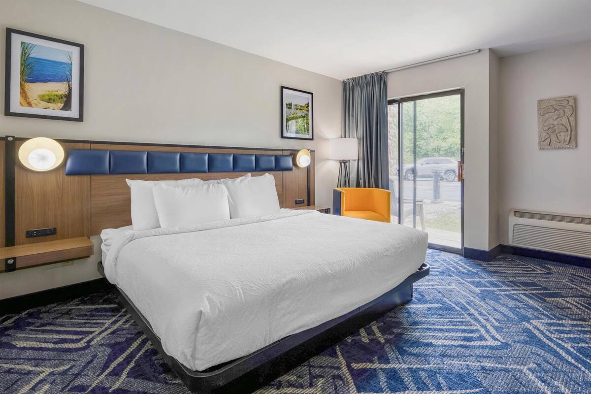 A hotel room with a large bed, a bedside table with a lamp, a window with curtains, and artwork on the walls.
