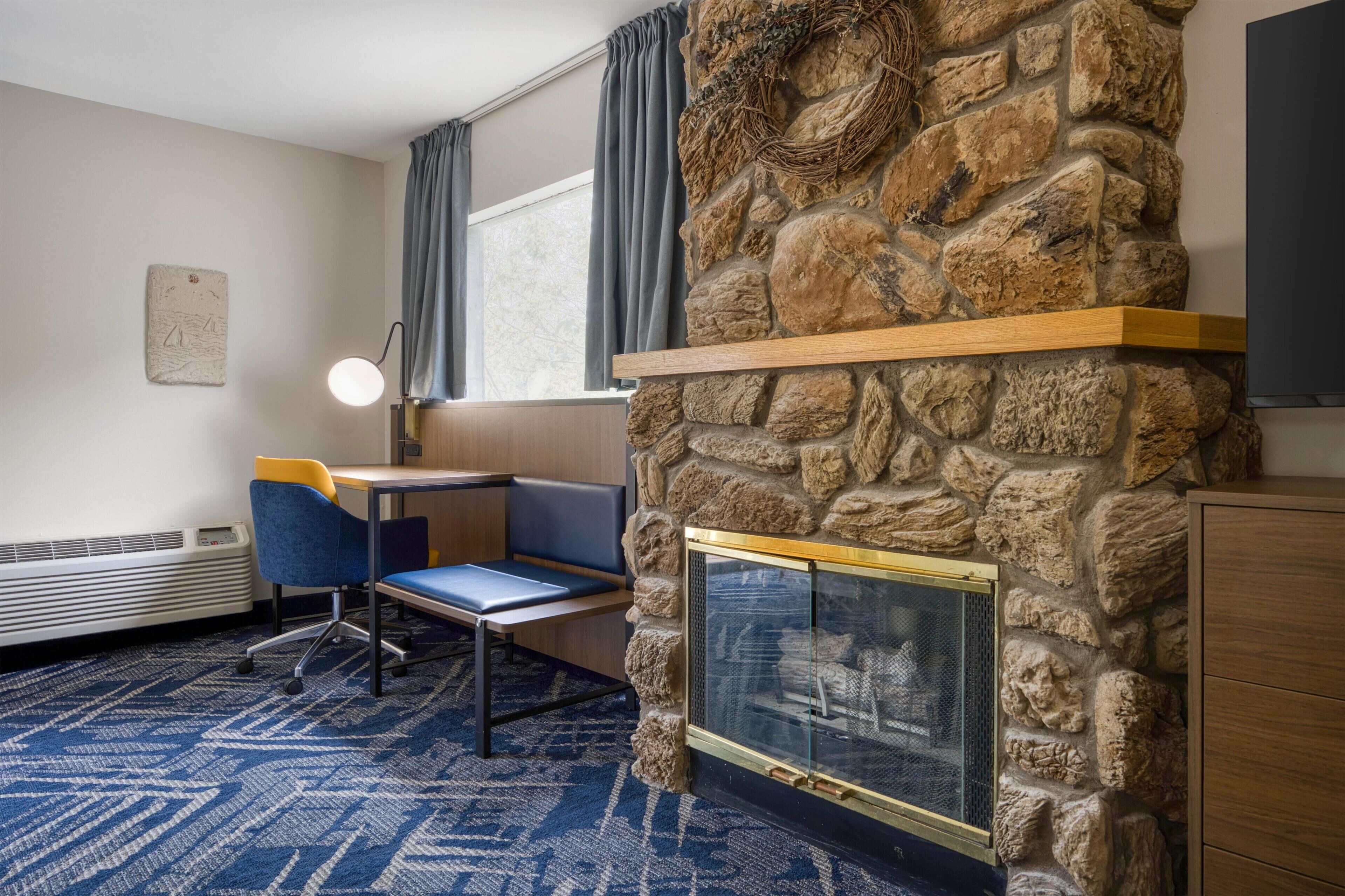 A room with a stone fireplace, a desk with a chair, a blue patterned carpet, and a window with curtains.