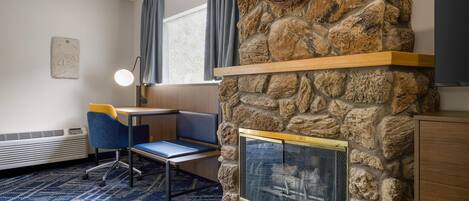 A room with a stone fireplace, a desk with a chair, a blue patterned carpet, and a window with curtains.