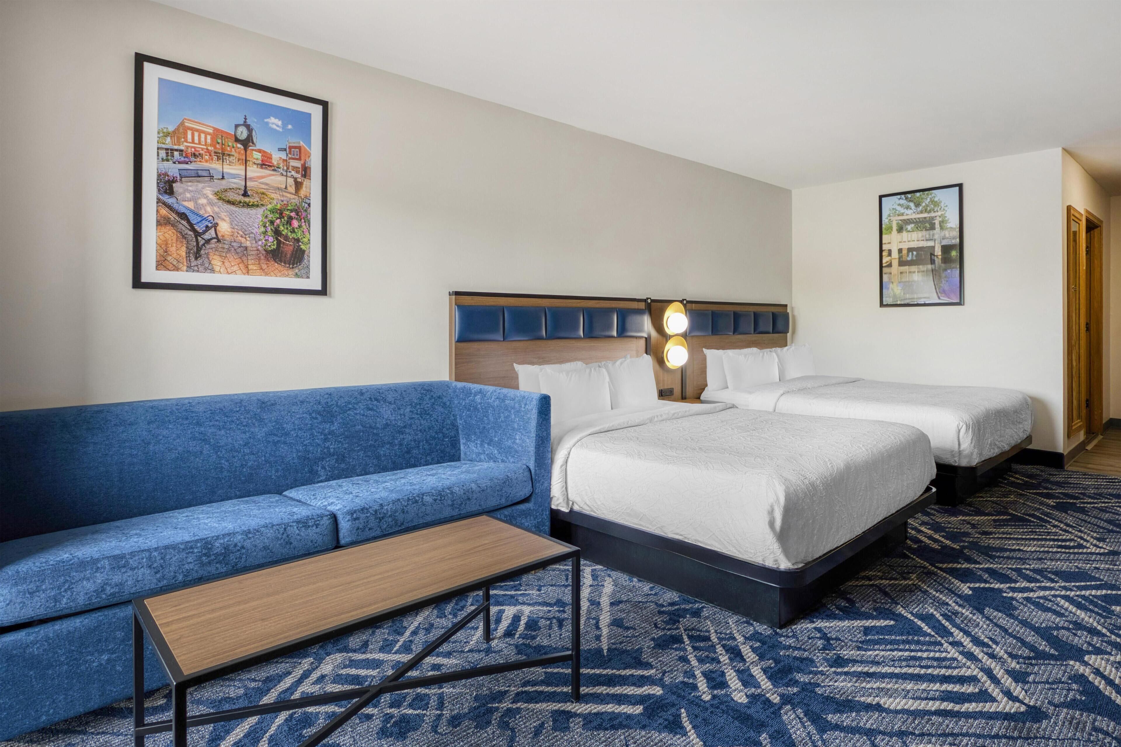 A hotel room with a blue sofa, a wooden coffee table, two beds with white bedding, and a patterned rug.