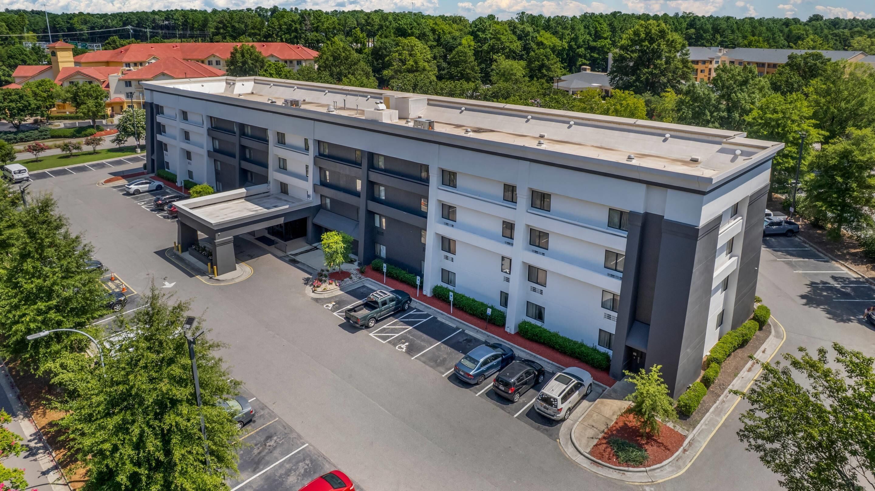 A multi-story hotel with a flat roof, parking lot, and surrounding greenery.