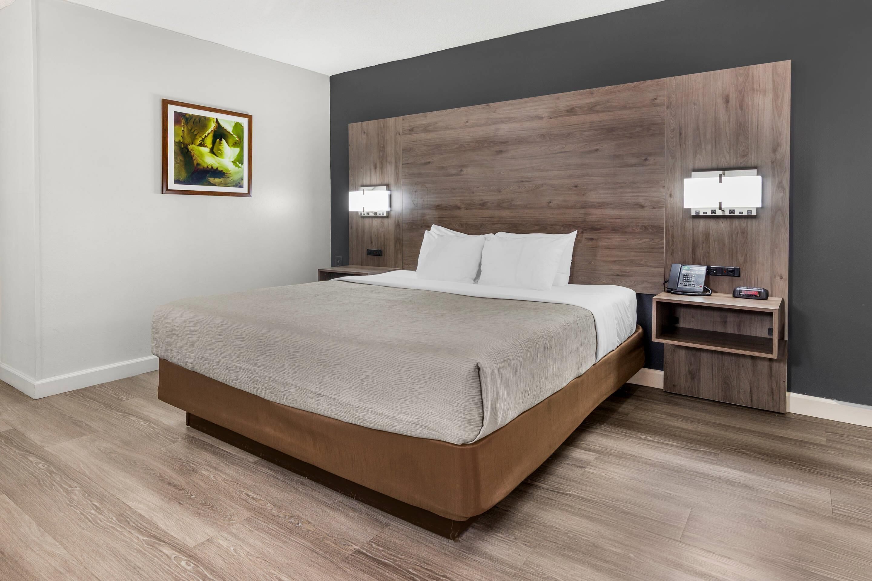 A modern hotel room with a large bed, wooden headboard, bedside lamps, and a nightstand with a phone and a small table.