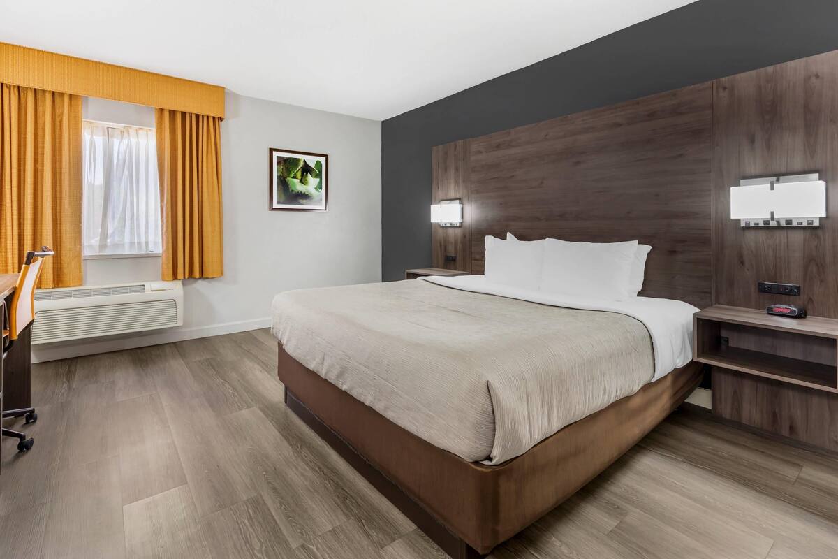 Une chambre d’hôtel avec un grand lit, une tête de lit en bois, deux lampes de chevet, une table de chevet, un bureau avec une chaise et une fenêtre avec des rideaux.