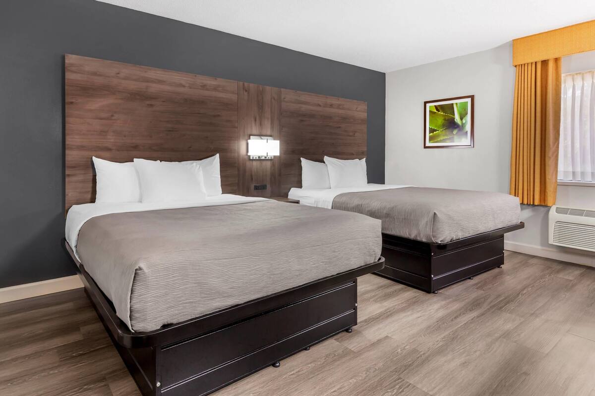 Une chambre d’hôtel avec deux lits, une tête de lit en bois, une lampe fixée au mur et une fenêtre avec des rideaux.