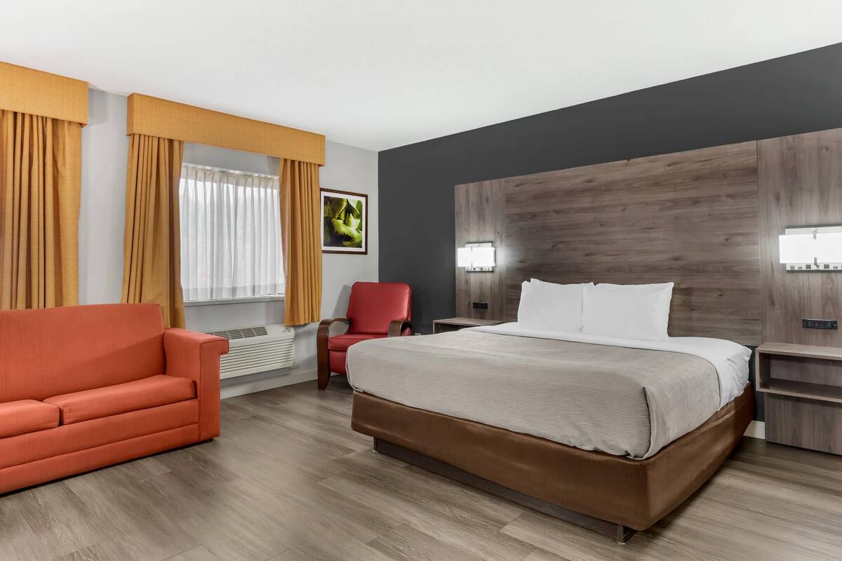 Une chambre d’hôtel moderne avec un grand lit, un fauteuil rouge, une tête de lit en bois et une grande fenêtre avec des rideaux.