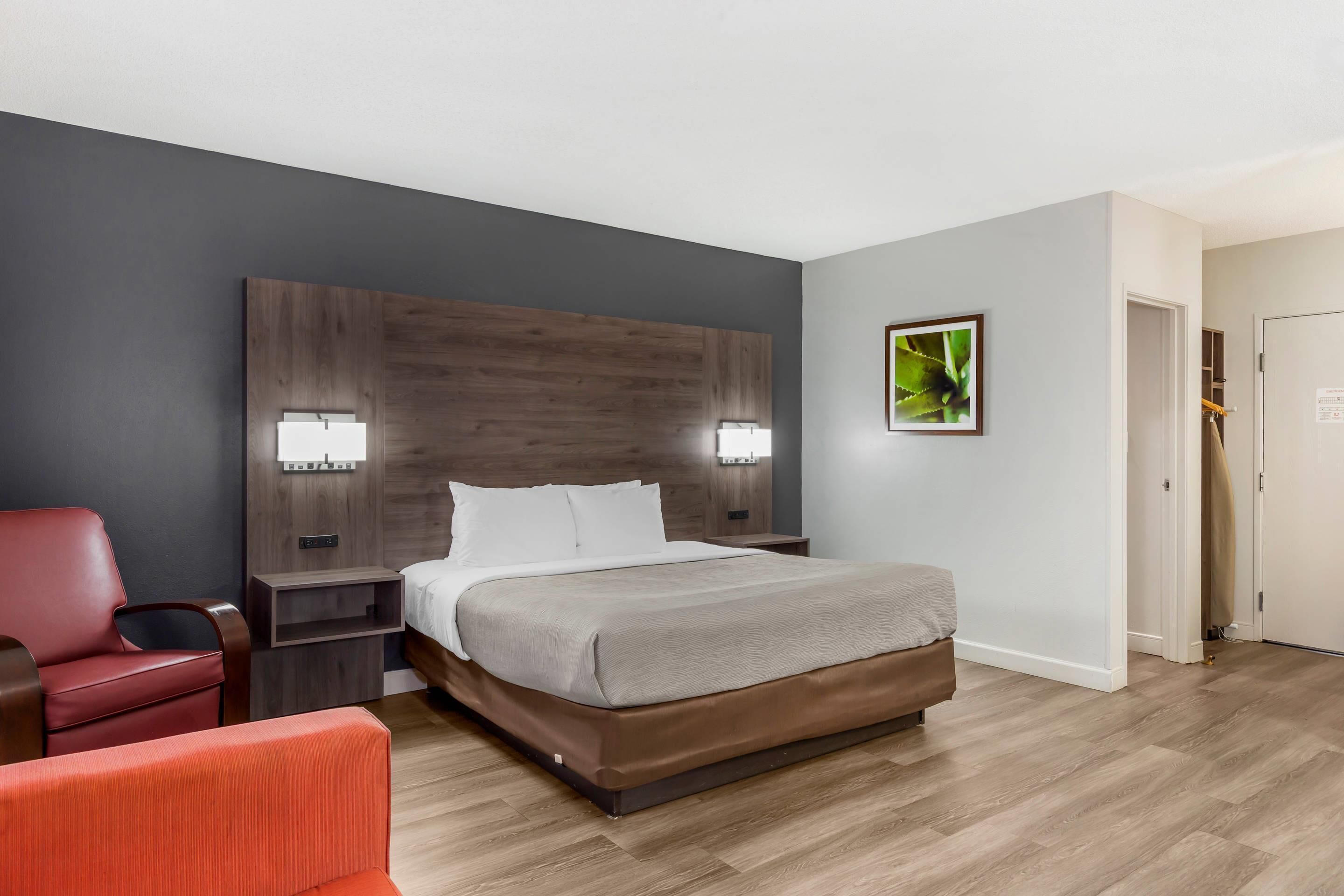 Una habitación de hotel con una cama grande, dos mesitas de noche con lámparas, un sillón rojo y un cabecero de madera.
