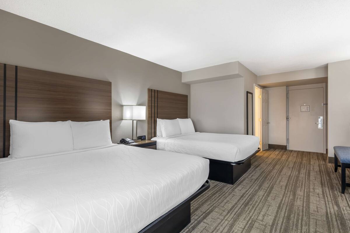 A hotel room with two beds, a nightstand, a lamp, and a bench.