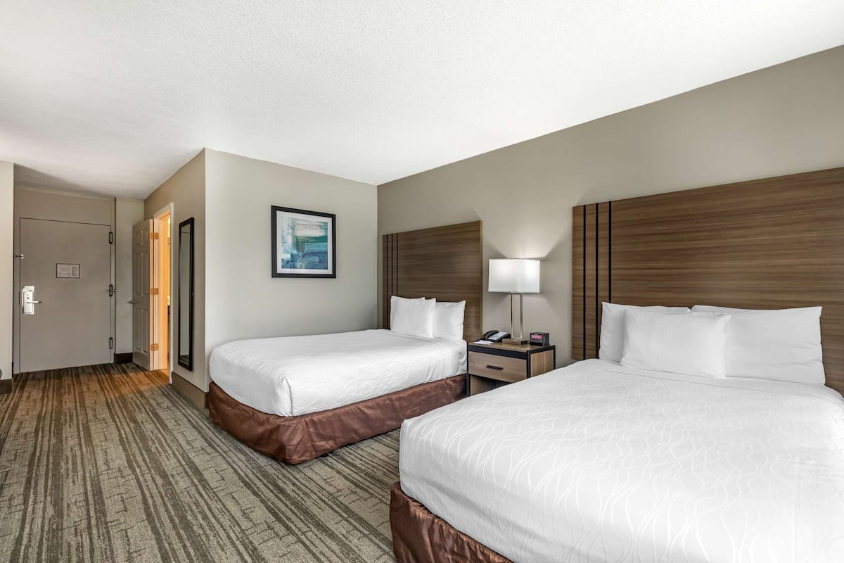 A hotel room with two beds, a wooden headboard, a bedside table with a lamp, and a framed picture on the wall.