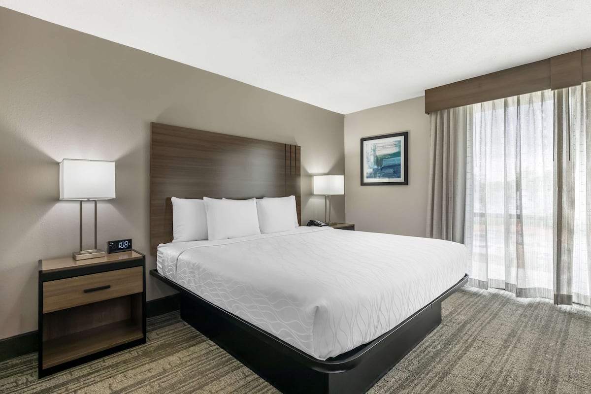 A hotel room with a large bed, two bedside tables with lamps, a headboard, a framed picture on the wall, and a window with curtains.
