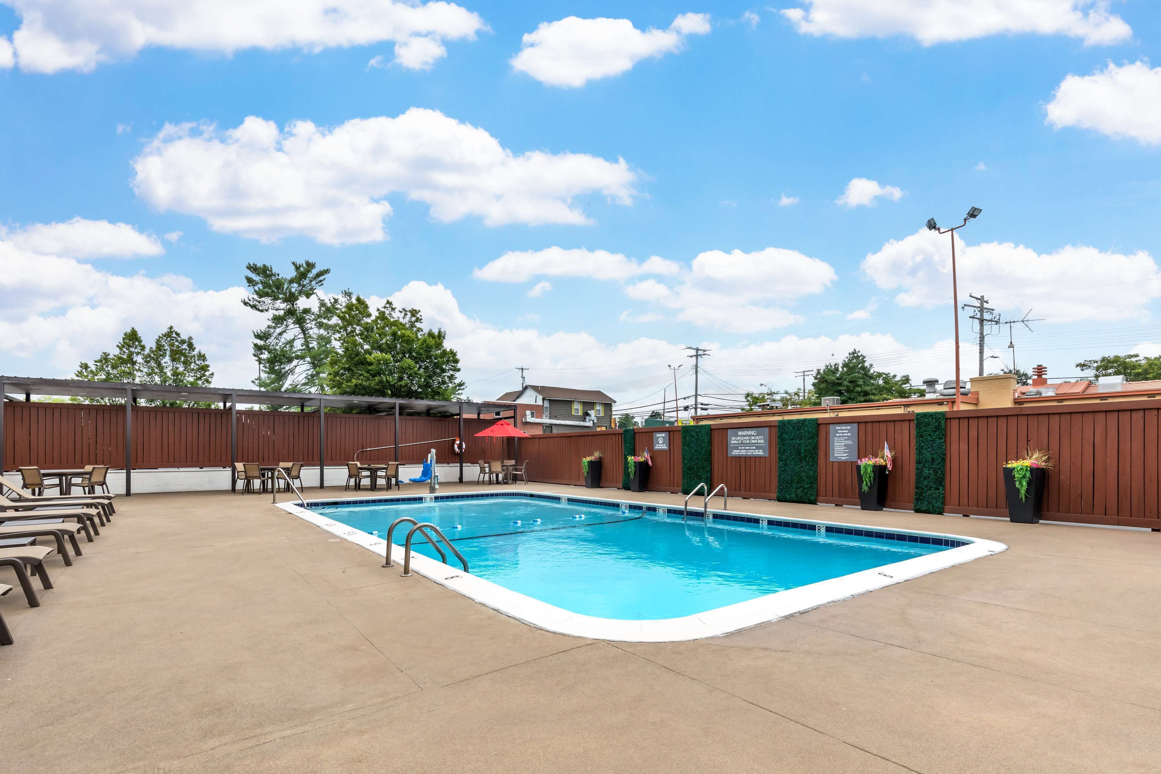 Una piscina rectangular con revestimiento azul, rodeada por una terraza de concreto. Se observan sillones de descanso y un paraguas rojo.