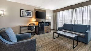 Habitación de hotel moderna con sofá, sillón, escritorio, televisión y un amplio ventanal con cortinas.