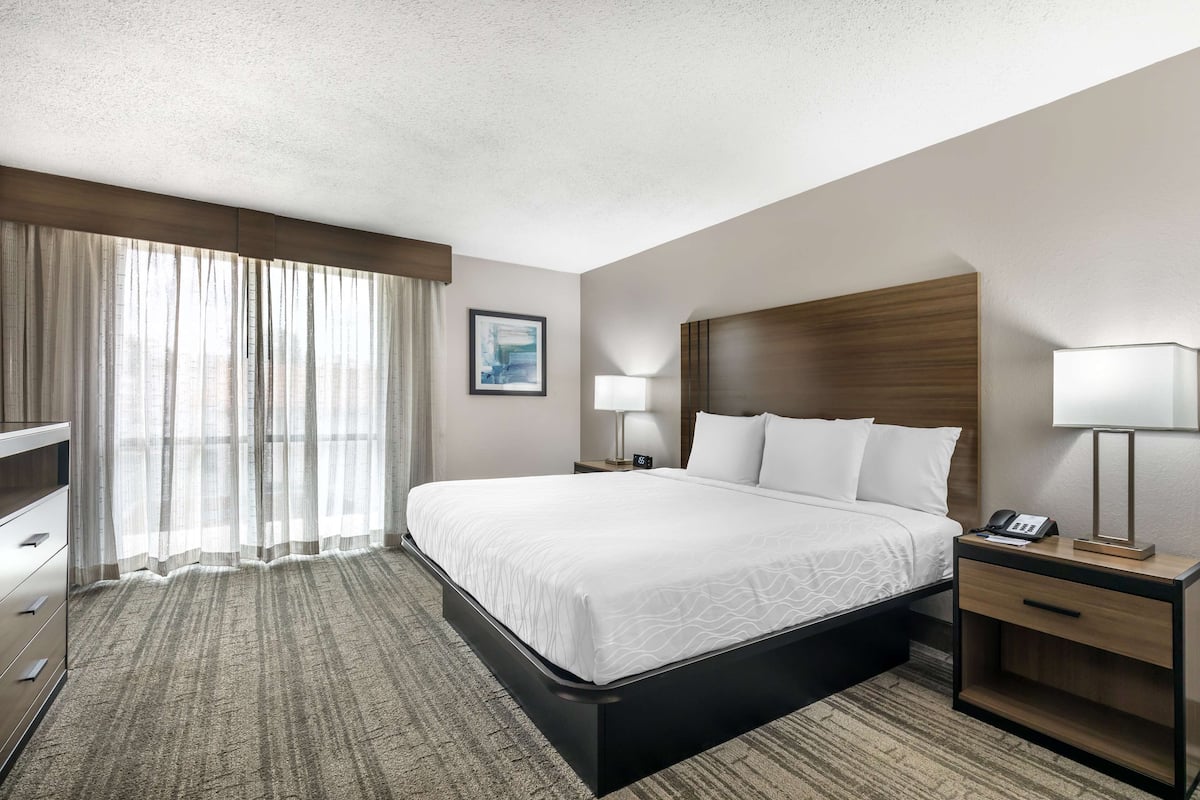 A hotel room with a large bed, a nightstand with a lamp, a desk with a telephone, and a view of the city through sheer curtains.
