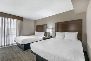 A hotel room with two beds, a large window with sheer curtains, a bedside table with a lamp, and a headboard with a wooden panel design.