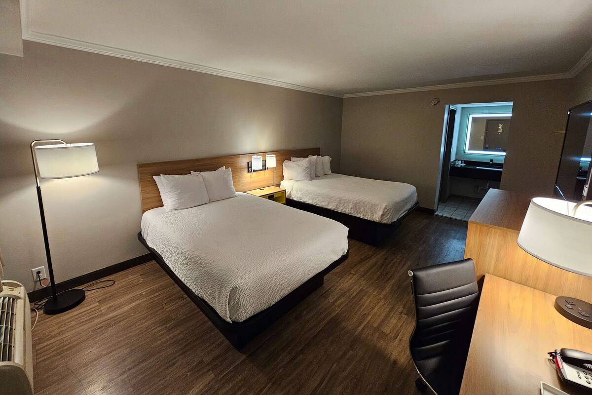 A hotel room with two beds, a desk, a chair, a lamp, and a small bathroom visible through an open door.