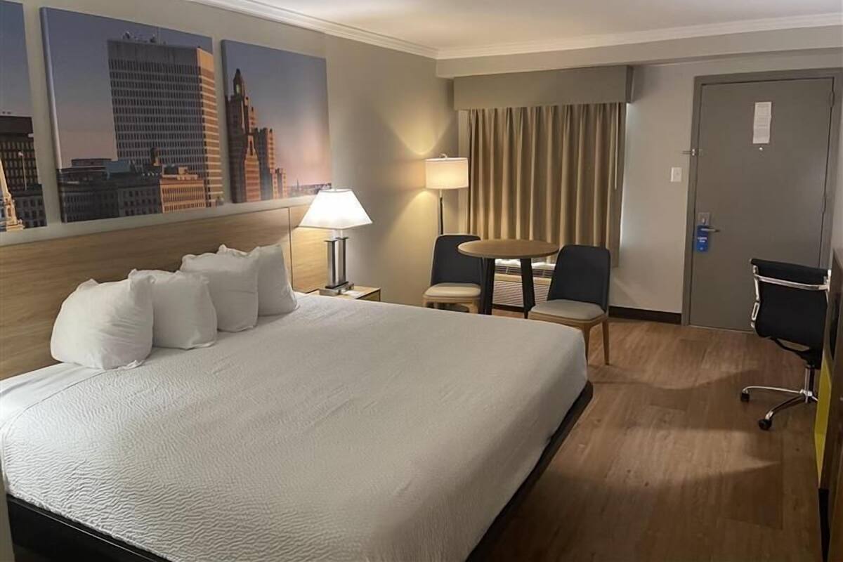 A hotel room with a large bed, a desk, two chairs, a lamp, and a view of city buildings.