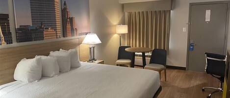 A hotel room with a large bed, a desk, two chairs, a lamp, and a view of city buildings.