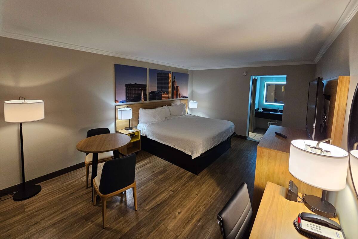 A hotel room with a bed, bedside tables, a desk, a chair, and a cityscape view.
