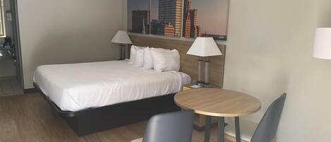 A hotel room with a bed, two chairs, a small table, and a cityscape painting on the wall.