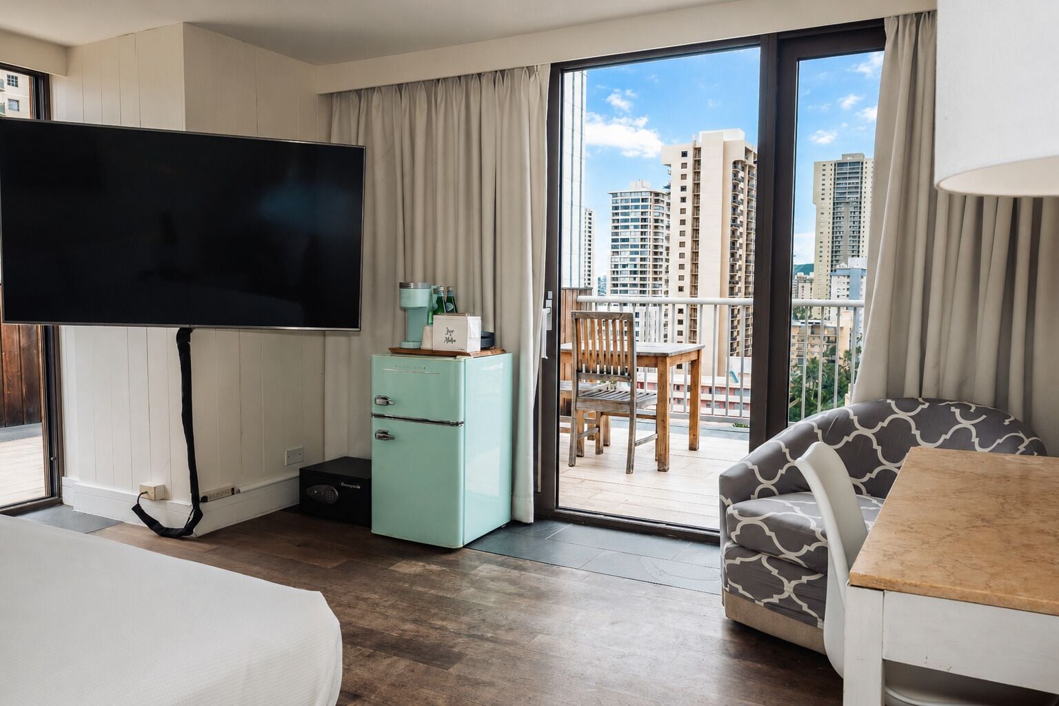 A hotel room with a large TV, a minibar, a balcony with a view of tall buildings, and a sofa.