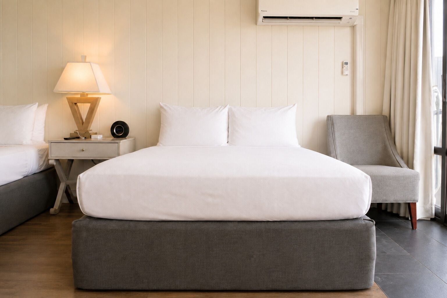 A hotel room with a bed, a bedside table with a lamp, a chair, and an air conditioning unit.
