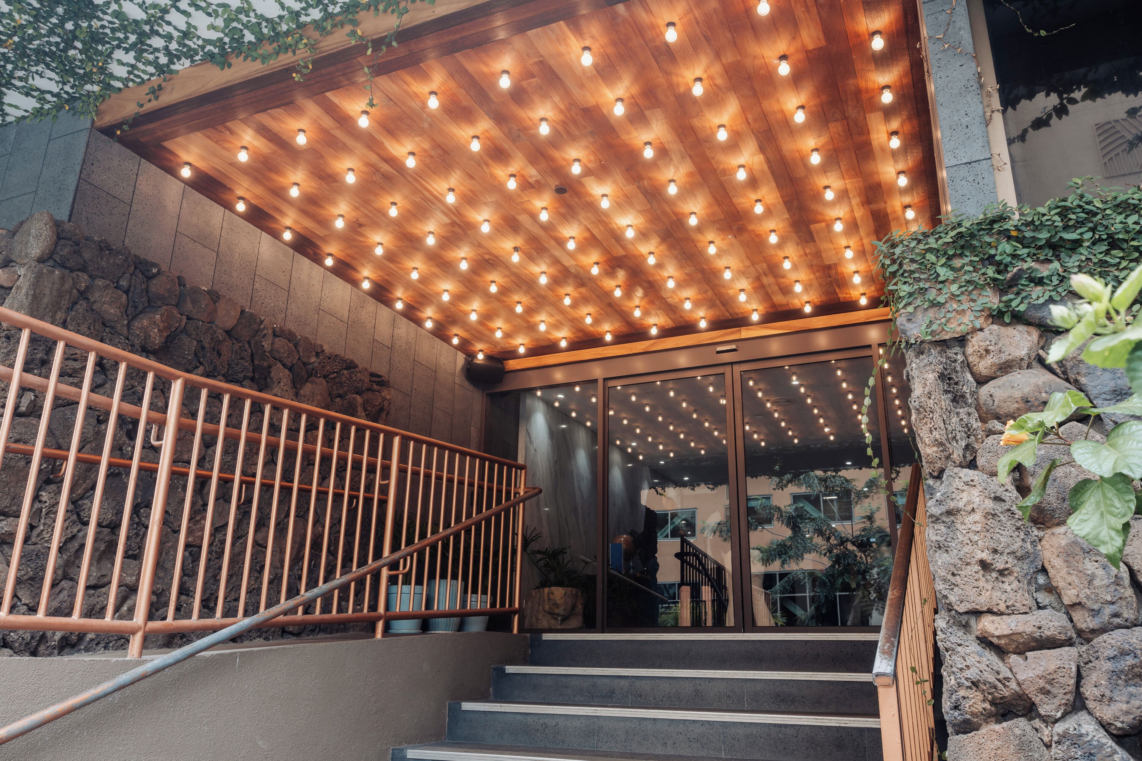 Una scala con ringhiera in metallo conduce all'ingresso di un edificio con soffitto in legno e luci decorative.