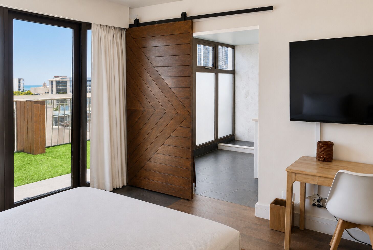 A hotel room with a bed, a desk, a chair, a TV, a sliding door, and a view of the city.