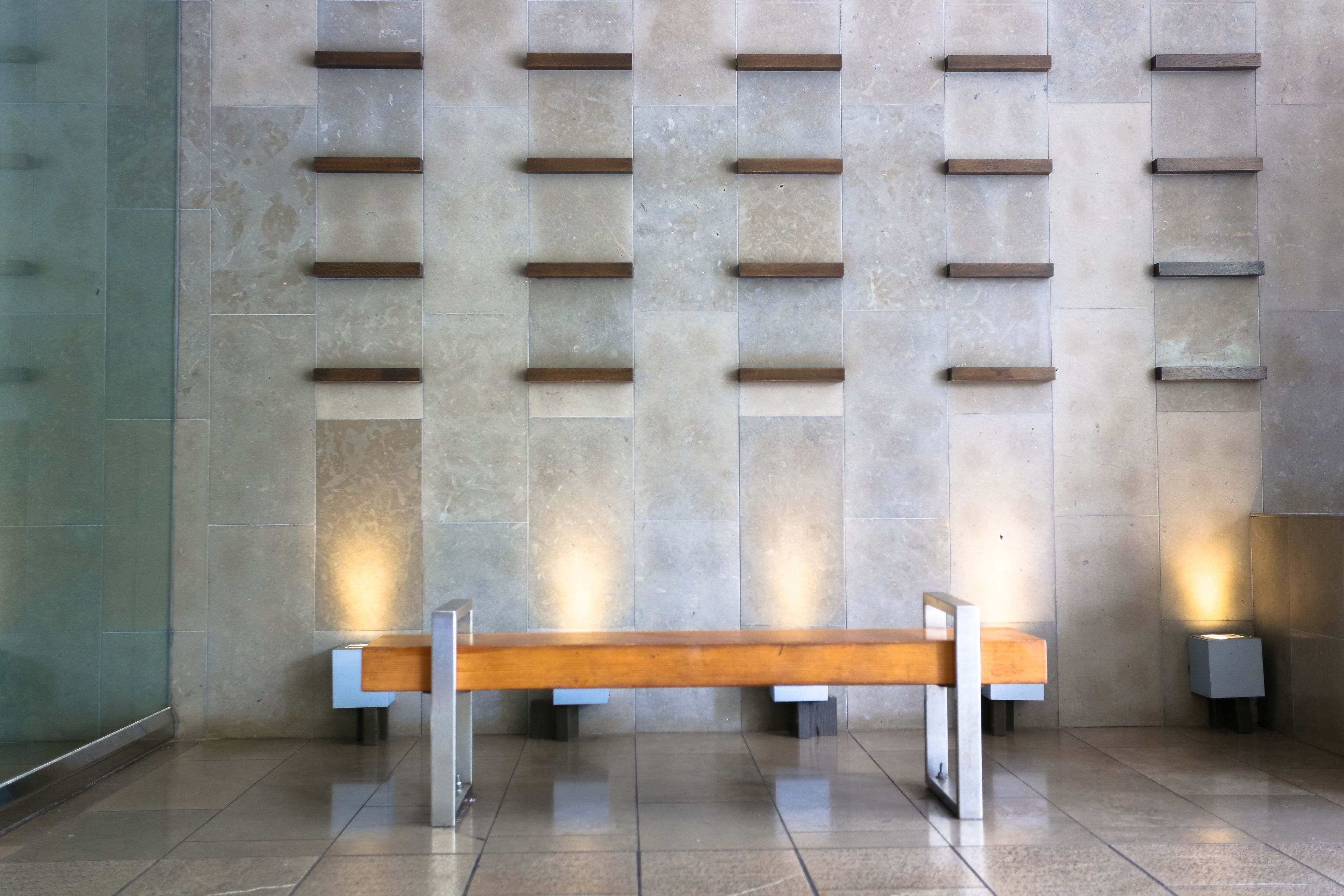 A modern bench with a wooden seat and metal frame, positioned in front of a wall with evenly spaced, rectangular cutouts.