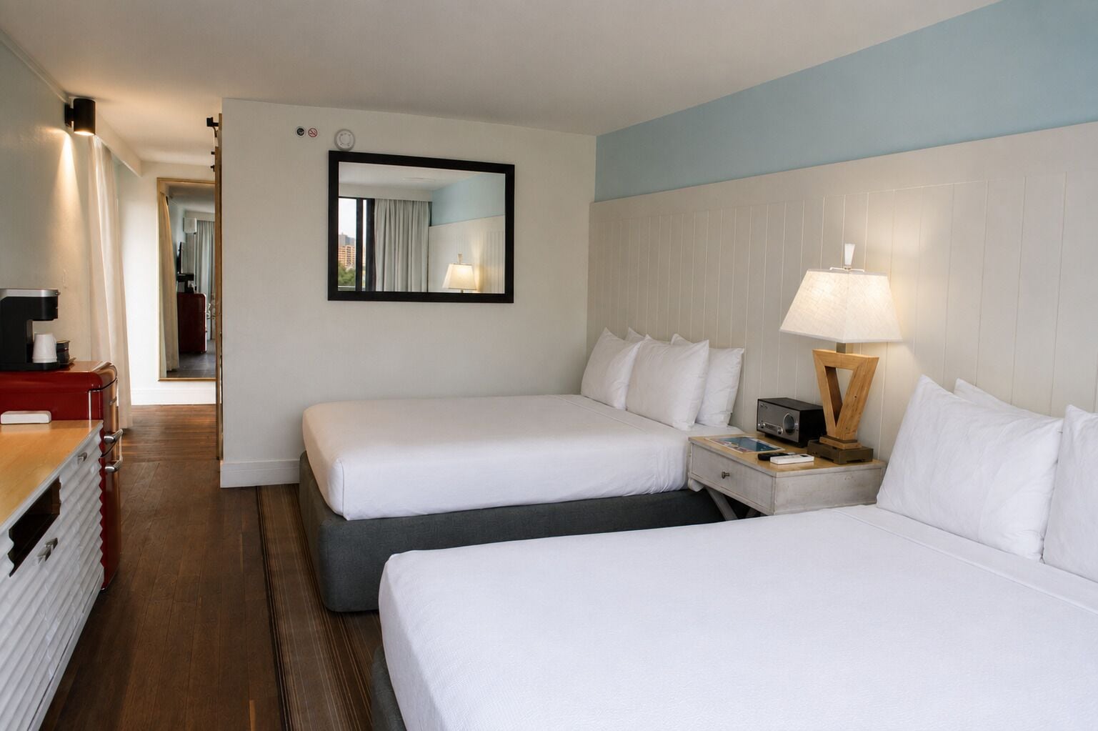 A hotel room with two beds, a desk, a lamp, and a mirror.