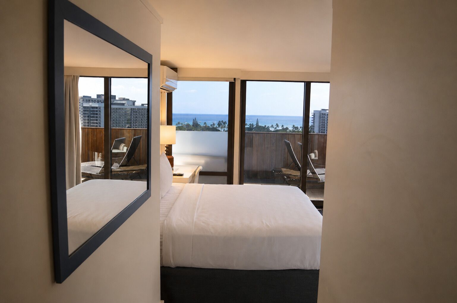 A hotel room with a bed, a desk, and a chair. There is a large window with a view of buildings and the ocean.