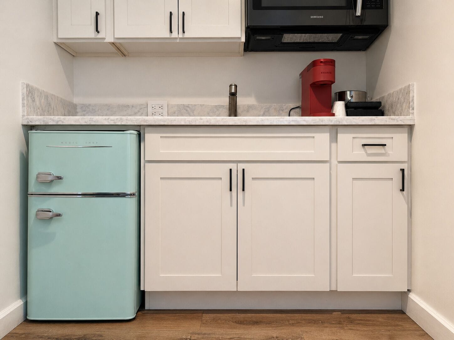 Une cuisine avec des meubles blancs, un réfrigérateur de couleur turquoise, un four à micro-ondes noir et une machine à café rouge.
