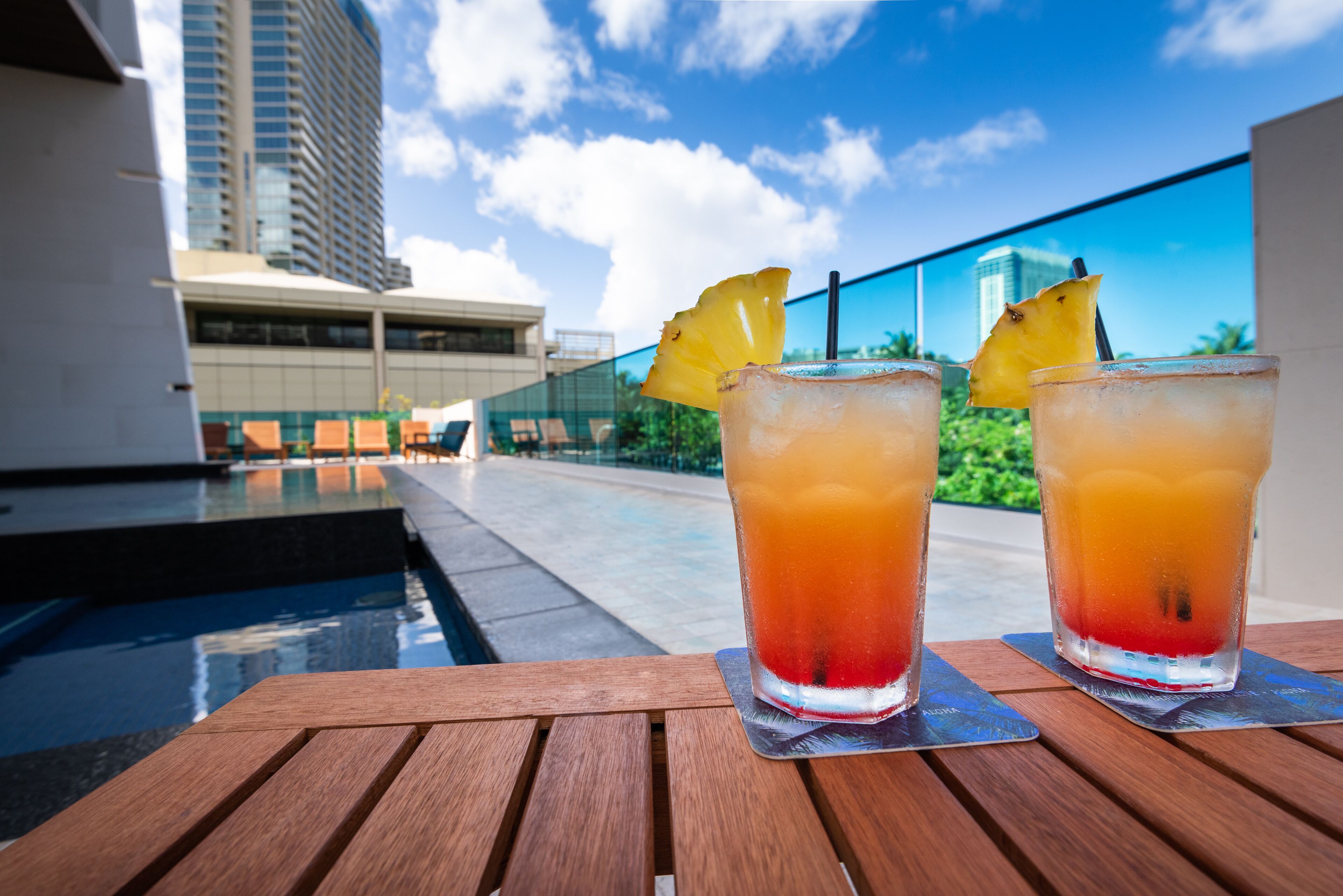 Dos vasos de una bebida tropical con una guarnición de piña sobre una mesa de madera junto a una piscina, con vistas a un edificio moderno y un cielo despejado.
