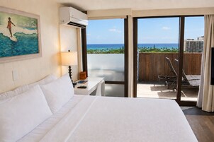 A hotel room with a bed, a desk, a chair, a painting, an air conditioning unit, and a balcony with a view of the ocean and palm trees.