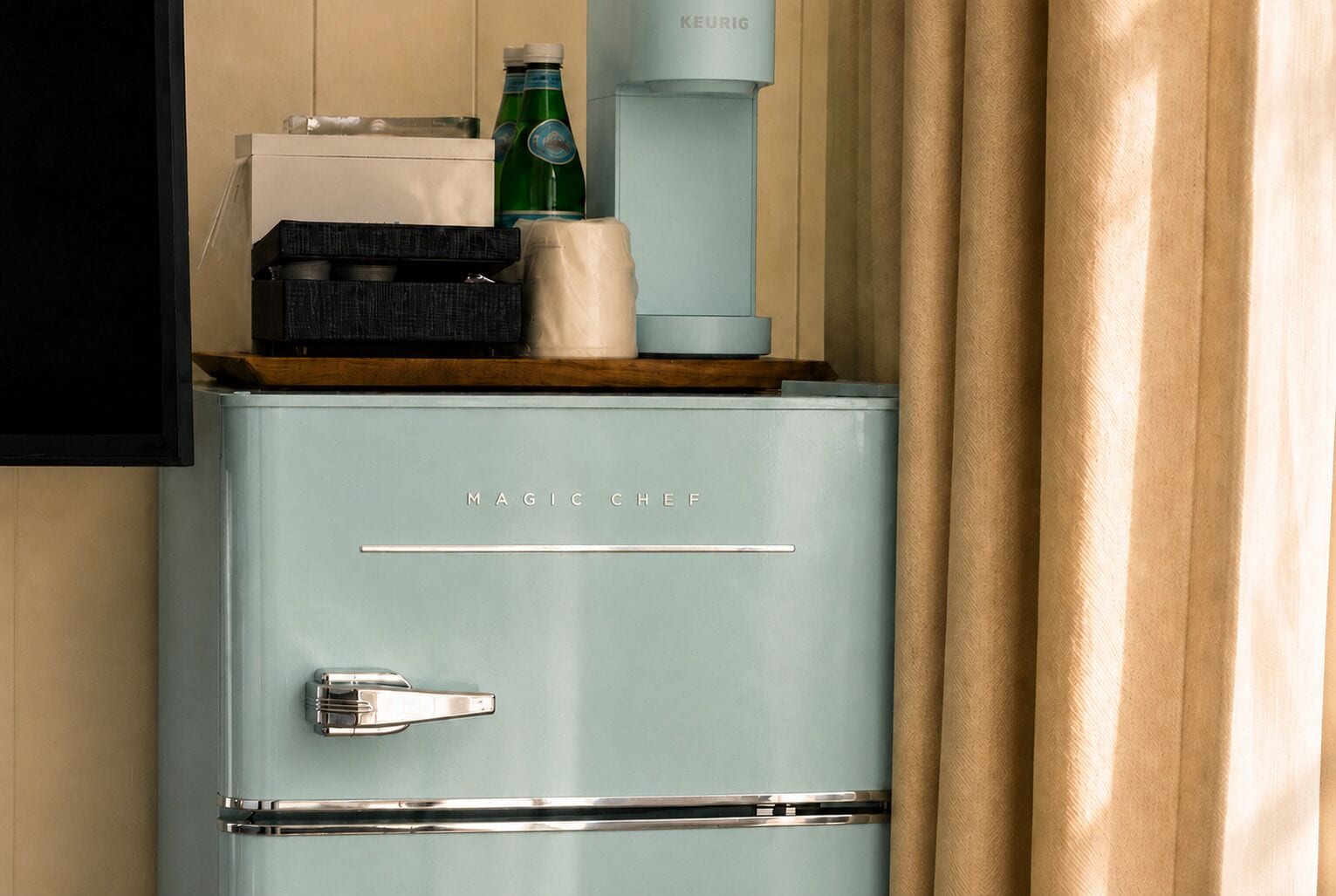 A vintage-style refrigerator with the brand name 'MAGIC CHEF' on it, placed in a room with beige walls and a curtain.