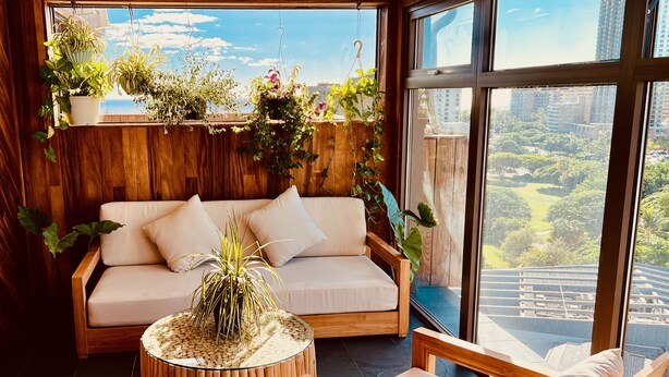 A modern living room with a sofa, a round wooden coffee table, and a chair. There are multiple hanging plants and a view of a cityscape through large windows. - Hotel La Croix (Honolulu)