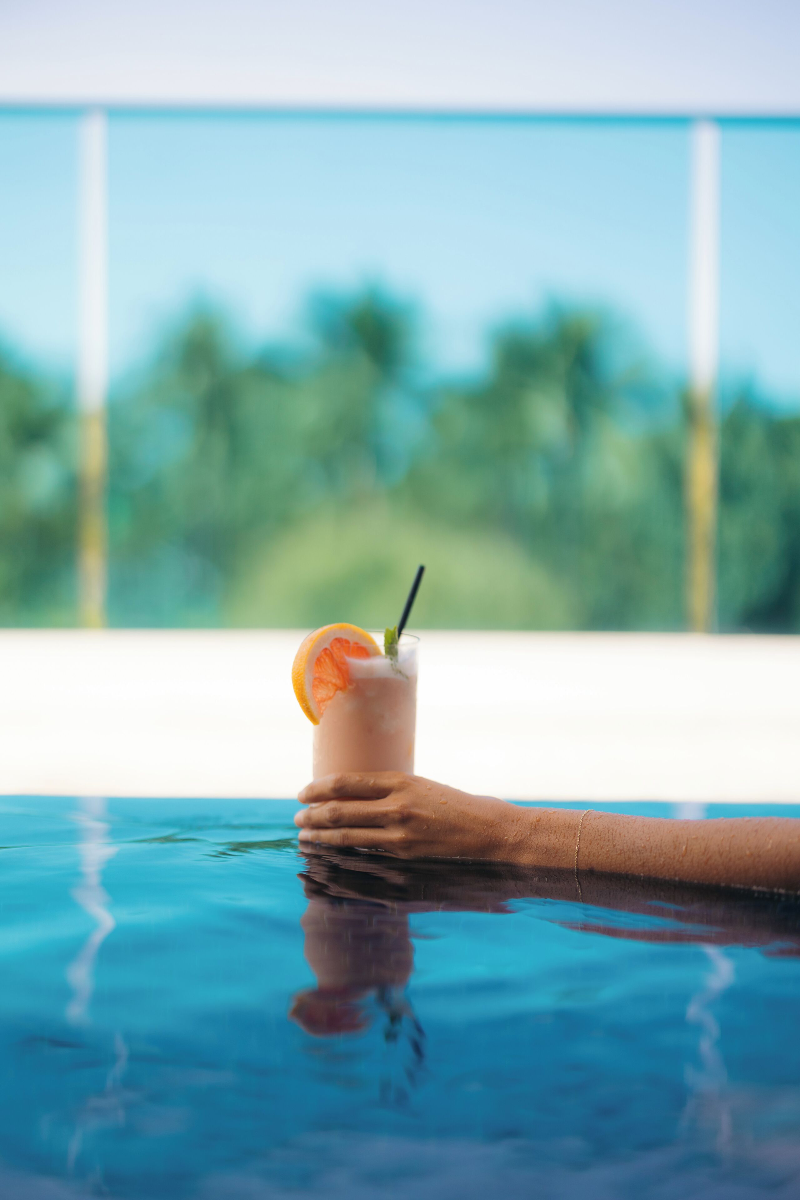 Una mano sosteniendo una bebida con pajita y una rodaja de fruta sobre una piscina.