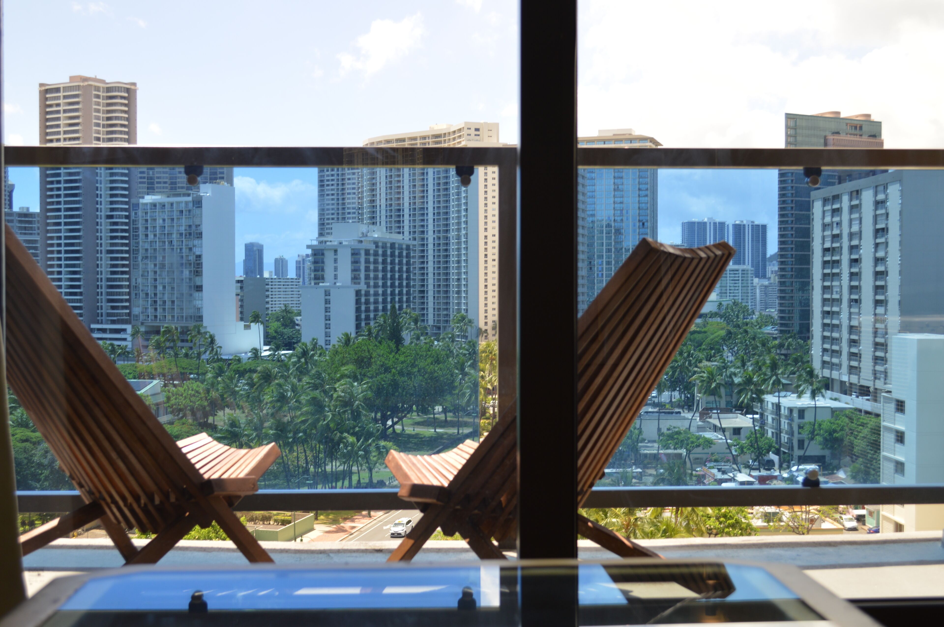 A view from a balcony with two wooden chairs, overlooking a cityscape with tall buildings and palm trees.